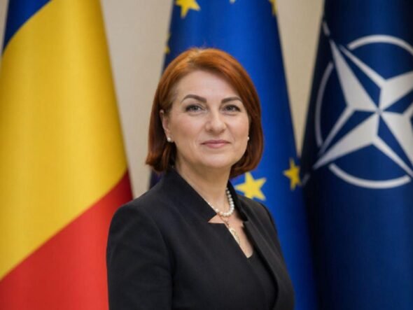 Female official posing in front of European Union and NATO flags, wearing a dark blazer and pearl necklace.