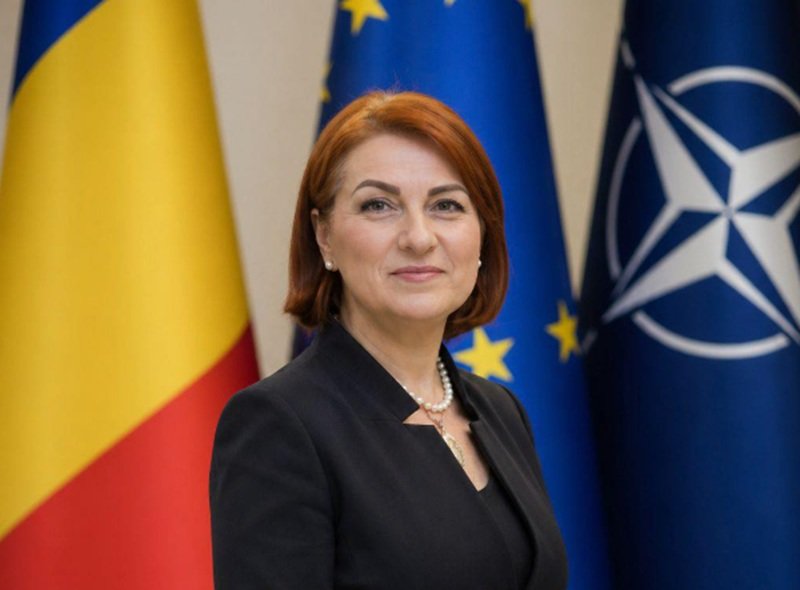 Female official posing in front of European Union and NATO flags, wearing a dark blazer and pearl necklace.