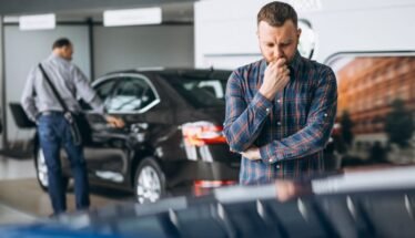 Man in a plaid shirt stands in a car showroom, deep in thought, with a black car and another person in the background near the display one.