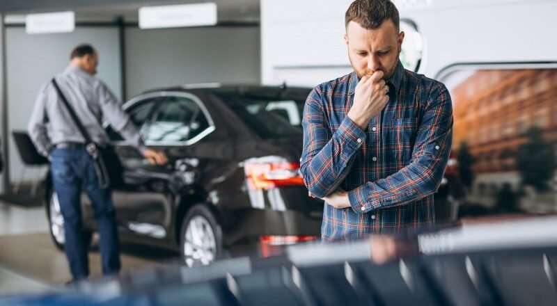 Man in a plaid shirt stands in a car showroom, deep in thought, with a black car and another person in the background near the display one.