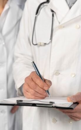 Two doctors in white coats; one writes on a clipboard while the other stands beside, stethoscope around the neck.