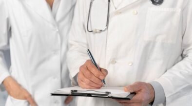 Two doctors in white coats; one writes on a clipboard while the other stands beside, stethoscope around the neck.