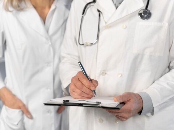 Two doctors in white coats; one writes on a clipboard while the other stands beside, stethoscope around the neck.