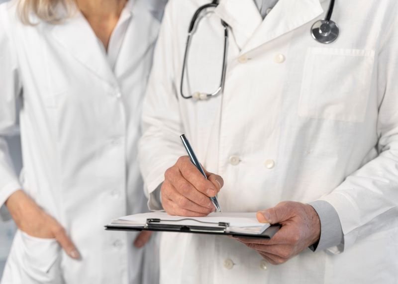 Two doctors in white coats; one writes on a clipboard while the other stands beside, stethoscope around the neck.