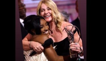Two women hug on stage at an awards ceremony, smiling, with a crystal trophy visible.