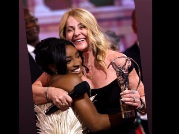 Two women hug on stage at an awards ceremony, smiling, with a crystal trophy visible.