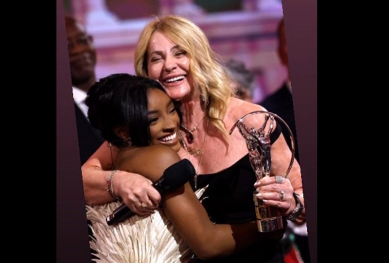 Two women hug on stage at an awards ceremony, smiling, with a crystal trophy visible.