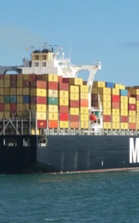 Container ship loaded with multicolored shipping containers, sailing on calm sea beneath a blue sky (MSC logo on hull).