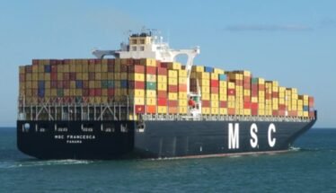 Container ship loaded with multicolored shipping containers, sailing on calm sea beneath a blue sky (MSC logo on hull).