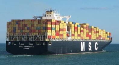 Container ship loaded with multicolored shipping containers, sailing on calm sea beneath a blue sky (MSC logo on hull).