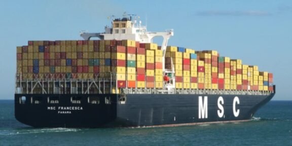 Container ship loaded with multicolored shipping containers, sailing on calm sea beneath a blue sky (MSC logo on hull).
