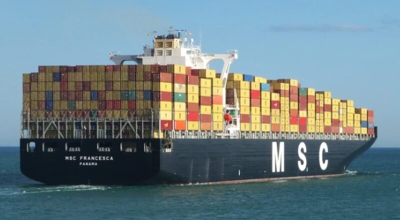 Container ship loaded with multicolored shipping containers, sailing on calm sea beneath a blue sky (MSC logo on hull).
