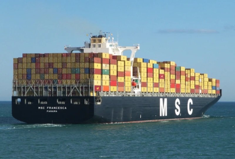 Container ship loaded with multicolored shipping containers, sailing on calm sea beneath a blue sky (MSC logo on hull).