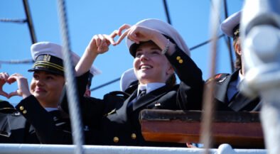 Cadets in navy uniforms on a ship deck making heart shapes with their hands against a clear blue sky.