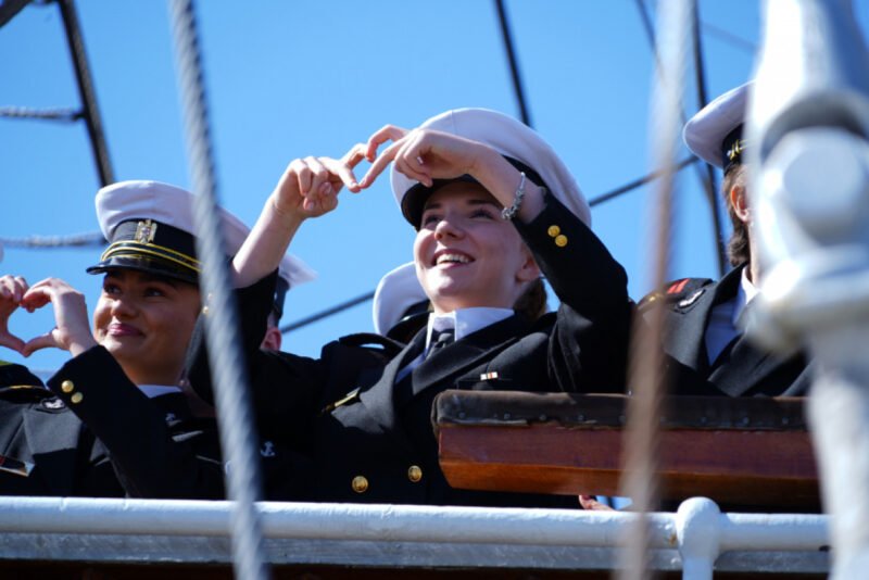 Cadets in navy uniforms on a ship deck making heart shapes with their hands against a clear blue sky.