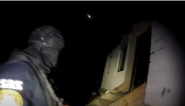 Rescue worker in dark gear and helmet standing beside a partially collapsed house at night, illuminated by a flashlight