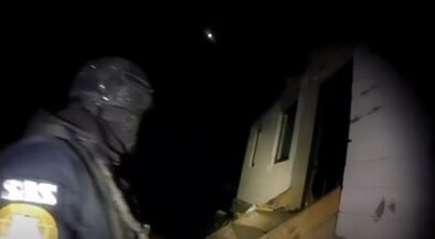 Rescue worker in dark gear and helmet standing beside a partially collapsed house at night, illuminated by a flashlight