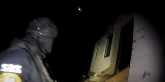 Rescue worker in dark gear and helmet standing beside a partially collapsed house at night, illuminated by a flashlight