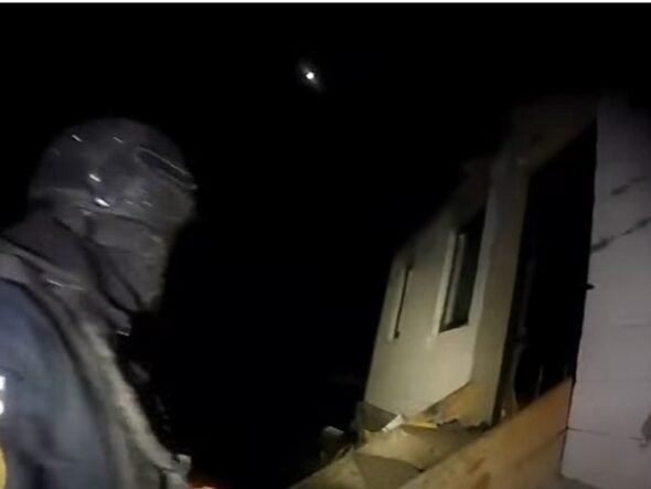 Rescue worker in dark gear and helmet standing beside a partially collapsed house at night, illuminated by a flashlight