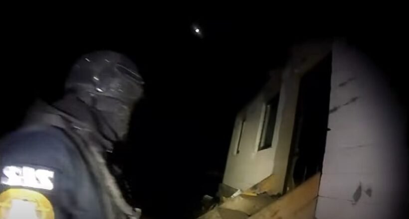 Rescue worker in dark gear and helmet standing beside a partially collapsed house at night, illuminated by a flashlight