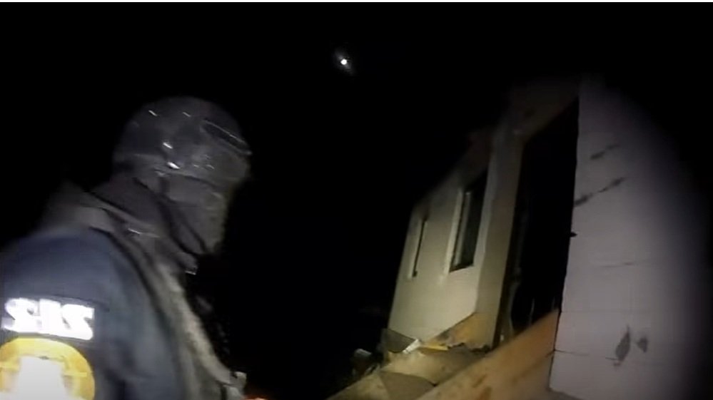 Rescue worker in dark gear and helmet standing beside a partially collapsed house at night, illuminated by a flashlight