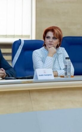 Three professionals seated at a curved conference table during a formal meeting; center woman with red hair in a light blazer flanked by two men in suits.