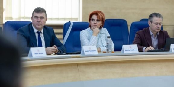 Three professionals seated at a curved conference table during a formal meeting; center woman with red hair in a light blazer flanked by two men in suits.