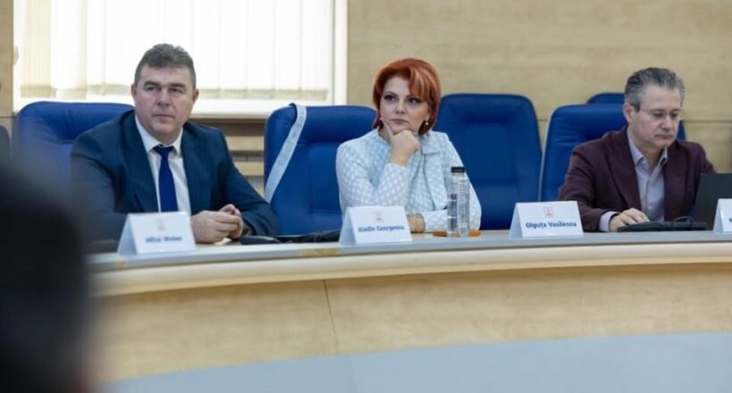 Three professionals seated at a curved conference table during a formal meeting; center woman with red hair in a light blazer flanked by two men in suits.