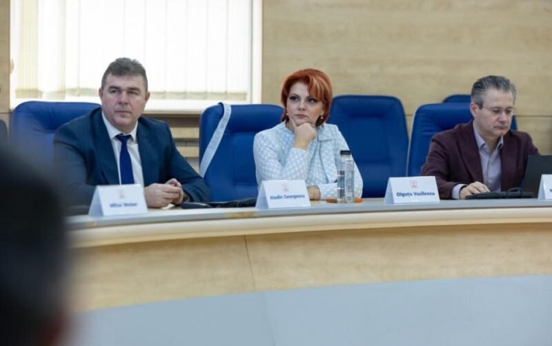 Three professionals seated at a curved conference table during a formal meeting; center woman with red hair in a light blazer flanked by two men in suits.