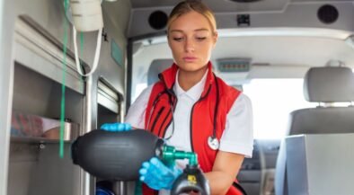Paramedic in a red vest with gloves and stethoscope treats a patient inside an ambulance, using a handheld medical device.