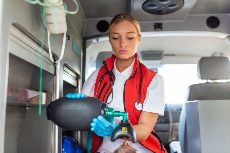 Paramedic in a red vest with gloves and stethoscope treats a patient inside an ambulance, using a handheld medical device.