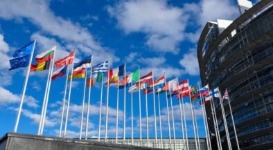 Row of colorful national flags on tall poles in front of a modern curved building against a blue sky outside an office complex.