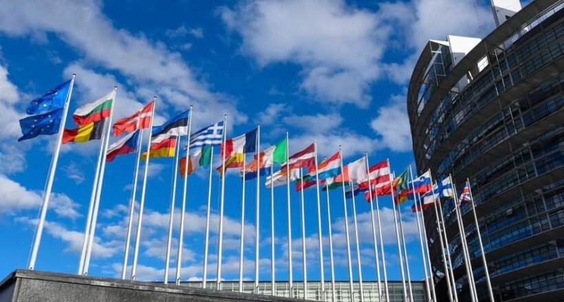 Row of colorful national flags on tall poles in front of a modern curved building against a blue sky outside an office complex.