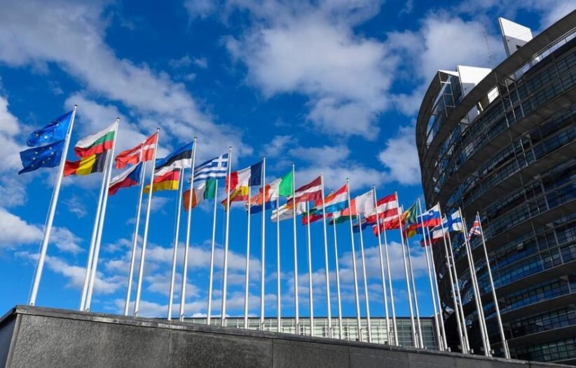 Row of colorful national flags on tall poles in front of a modern curved building against a blue sky outside an office complex.