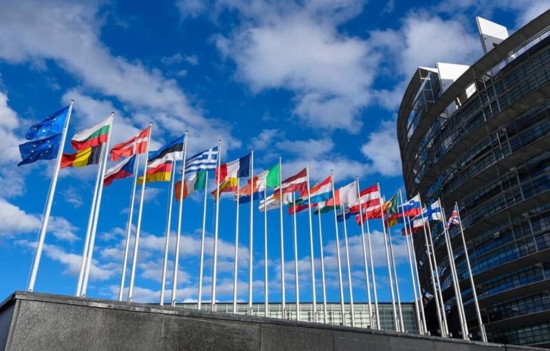 Row of colorful national flags on tall poles in front of a modern curved building against a blue sky outside an office complex.