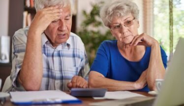 Elderly couple sitting at a table, looking worried while reviewing documents and a laptop.