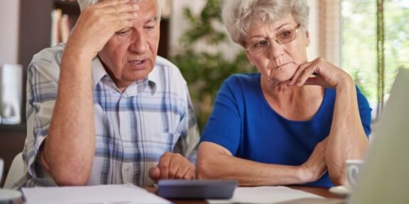 Elderly couple sitting at a table, looking worried while reviewing documents and a laptop.