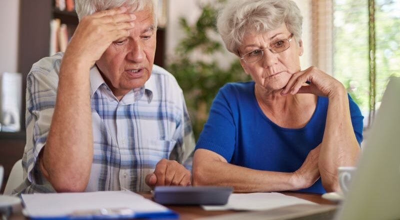 Elderly couple sitting at a table, looking worried while reviewing documents and a laptop.