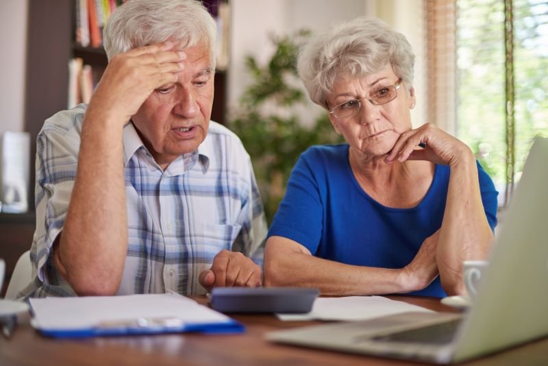 Elderly couple sitting at a table, looking worried while reviewing documents and a laptop.