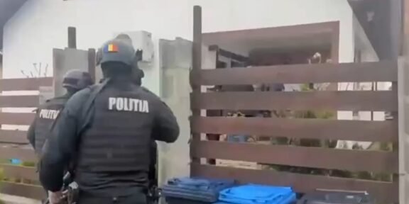 Two Romanian police officers in tactical gear approach a residential property gate, with blue recycling bins in the foreground.