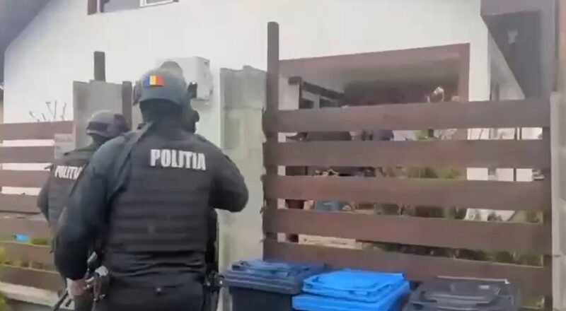 Two Romanian police officers in tactical gear approach a residential property gate, with blue recycling bins in the foreground.