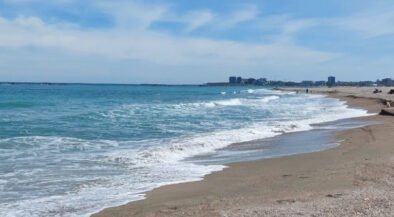 Sunny beach with gentle waves washing onto the sandy shore under a blue sky, distant city skyline on the horizon.]","Wide view of a turquoise sea meeting a sandy beach, with a distant coastal city row of buildings.