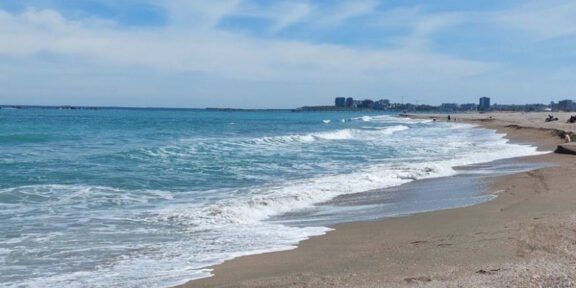 Sunny beach with gentle waves washing onto the sandy shore under a blue sky, distant city skyline on the horizon.]","Wide view of a turquoise sea meeting a sandy beach, with a distant coastal city row of buildings.
