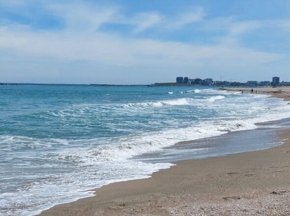 Sunny beach with gentle waves washing onto the sandy shore under a blue sky, distant city skyline on the horizon.]","Wide view of a turquoise sea meeting a sandy beach, with a distant coastal city row of buildings.