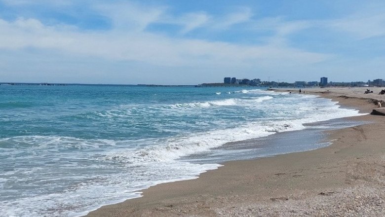 Sunny beach with gentle waves washing onto the sandy shore under a blue sky, distant city skyline on the horizon.]","Wide view of a turquoise sea meeting a sandy beach, with a distant coastal city row of buildings.