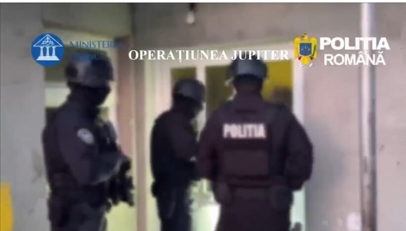 Armed Romanian police in tactical gear guard a doorway during Operation Jupiter (text overlay: OPERATIUNEA JUPITER).