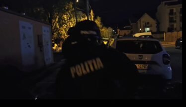 Nighttime street scene with a Romanian police officer wearing a jacket that reads POLITIA, standing near a white car on a dimly lit residential street.