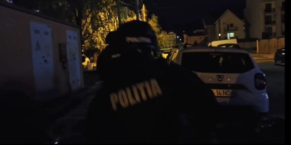 Nighttime street scene with a Romanian police officer wearing a jacket that reads POLITIA, standing near a white car on a dimly lit residential street.