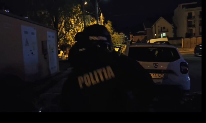 Nighttime street scene with a Romanian police officer wearing a jacket that reads POLITIA, standing near a white car on a dimly lit residential street.