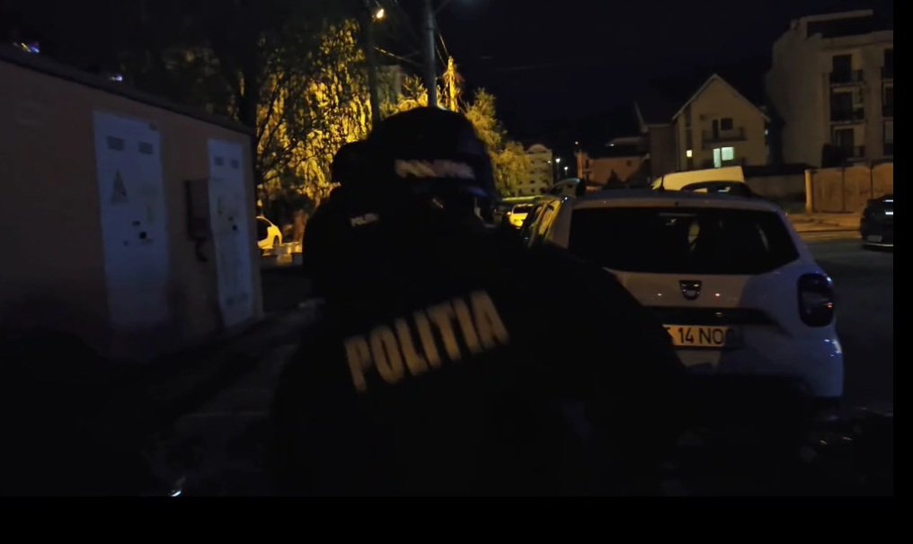 Nighttime street scene with a Romanian police officer wearing a jacket that reads POLITIA, standing near a white car on a dimly lit residential street.